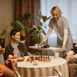 Group of senior Caucasian man and woman and Black woman sitting around table playing chess, while young adult Caucasian female caregiver standing nearby assisting in nursing home setting