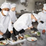 Multiracial group of cooks finishing main courses while working together in the kitchen. Cooks wearing uniform and face mask. Team prepares meals for the restaurant