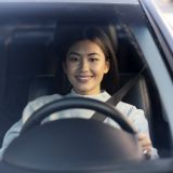 Asian smiling young woman enjoying driving on a beautiful day. Female driver sitting and holding steering wheel with pleasure smile