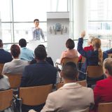 Rear view of Caucasian businessman raising hand while he is sitting in front of Asian businessman at business seminar in office building