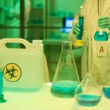 Contemporary clinician in coveralls and gloves holding two test tubes with blue liquid toxic substance while standing by workplace in lab
