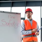 handsome firefighter in helmet holding clipboard and pen while standing near white board with fire