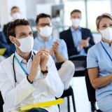 Male doctor and his colleagues wearing protective face masks and clapping their hands after successful education event in convention center.