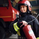 Holds extinguisher in hands. Female firefighter in protective uniform standing near truck.