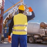 Back view of man engineer wearing work vest and safety helmet while looking at industrial equipment and truck at production plant
