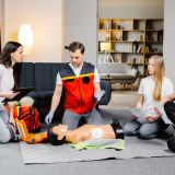 Young man instructor helping to make first aid heart compressions with dummy during the group training indoors.