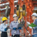 A team of engineers, including a female and two male professionals, conducts a safety inspection of lifting equipment in a metal sheet factory, ensuring compliance and operational efficiency.
