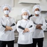 Portrait of multiracial team of three chefs standing together in the professional kitchen. Well-dressed chefs in face masks and protective gloves ready for a job. New normal for business during pandemic