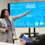 Young adult Hispanic woman presenting data on digital screen to Caucasian woman during business meeting, gesturing toward charts while colleague taking notes on tablet