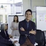 Young manager is smiling with arms crossed while attending a business meeting in a modern office with colleagues discussing in the background