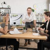 Caucasian businessman in white shirt smiling and showing financial report during conference with multiracial partners. Business people sitting at desk office with papers and modern technology.