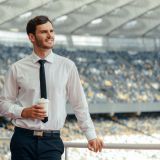 Man entrepreneur dressed in white shirt with tie, holding cup of coffee while standing leaning against the railing of observation deck of the stadium
