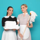 Young chambermaids with stack of clean towels and mop on color background