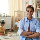 Biracial woman smiling at camera with arms crossed in modern nursery setting Child playing with toys in background and shelves filled with children's items