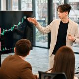 Young worker leading business meeting in an office, discussing the topic of cryptocurrencies with other workers using a big display with currency rate and physical coin