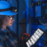 Side profile in a technical room with cool blue lighting, a specialist verifies a tablet schematic while inspecting wiring. Focused mood with smart maintenance and safety concept.